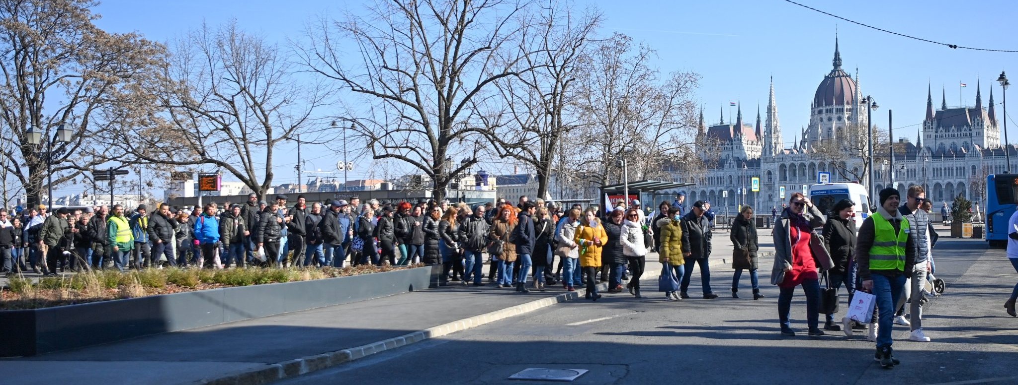 A demonstráció résztvevői a Batthyány térnél vonulnak, háttérben az Országház épületével.